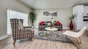 A living room with a plaid armchair, a white chair, a round coffee table, a grey sofa with red cushions, and a painting on the wall. - Modern 4BR/2B Home, with HotTub, Near DFW Airport (Irving)