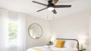 A bedroom with a bed, bedside tables, a ceiling fan, a window with curtains, and a wall-mounted mirror. - Modern 4BR/2B Home, with HotTub, Near DFW Airport (Irving)