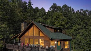 A two-story log cabin with large windows and a balcony, surrounded by dense forest.