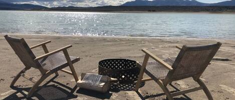 Two wicker chairs and a basket on a sandy beach with a body of water and mountains in the background.