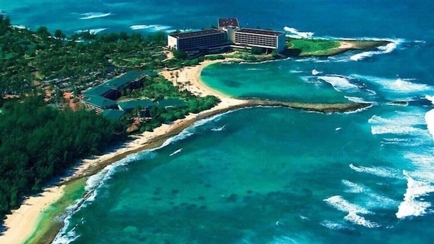 Vue aérienne d’un hôtel situé sur la côte, avec un grand bâtiment, une plage et les eaux environnantes.