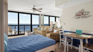 A bedroom with a large bed, a desk with chairs, a view of the ocean, and a ceiling fan.