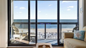 A room with a large sliding glass door leading to a beach view. Inside, there is a beige sofa with a starfish pillow and a wooden coffee table.