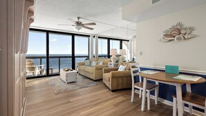A spacious living area with a large window overlooking the sea, a ceiling fan, and a dining table with chairs.