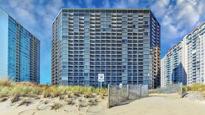 A high-rise building with a grid of windows, surrounded by sandy terrain and other buildings.