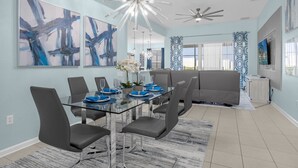 Un espace repas moderne avec une table en verre, des chaises grises et de la vaisselle bleue. Les murs sont ornés de peintures abstraites et le plafond est éclairé par un luminaire.