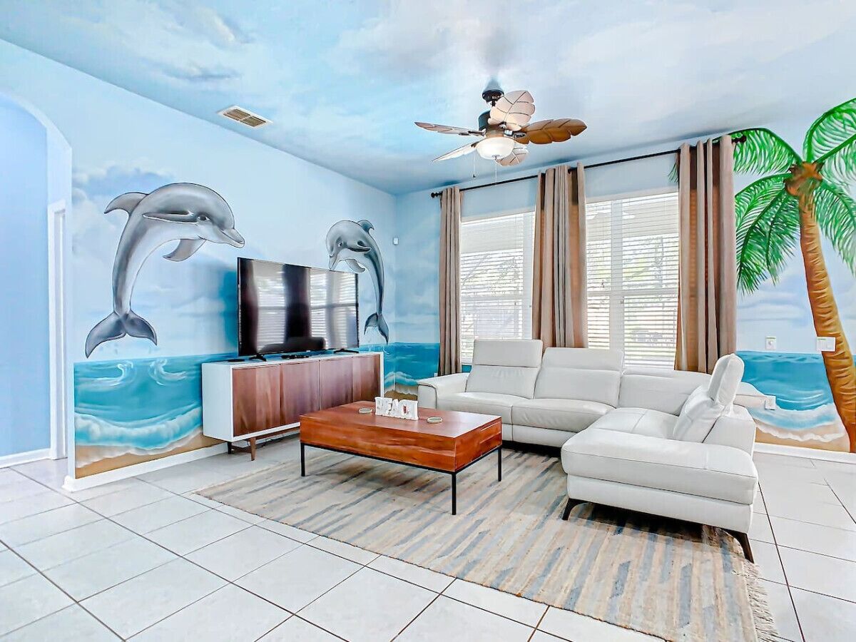 Una sala de estar con un gran mural de delfines, un ventilador de techo, un sofá blanco, una mesa de centro y un televisor.