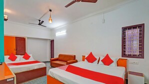 Quarto de hotel com duas camas, um sofá, um ventilador de teto e uma janela com grades.