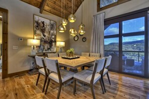 A dining area with a wooden table, six chairs, pendant lights, and a large window with a view.