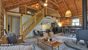 A cozy cabin interior with a wooden staircase, a living room with a sofa and coffee table, a dining area with chairs, and a kitchen with a stove. - Hygge House | Cozy Cabin + Fire Pit Near Downtown (Blue Ridge)