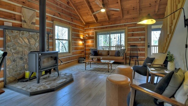 A log cabin interior with a stone fireplace, wooden furniture, and large windows. - Hygge House | Cozy Cabin + Fire Pit Near Downtown (Blue Ridge)