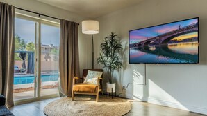 Un salón con un televisor grande, una silla, una planta y vista a una piscina.