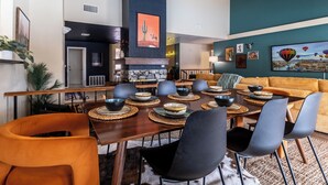 Un comedor con mesa de madera dispuesta para seis personas, rodeada de sillas negras. Hay un sofá amarillo y un televisor montado en la pared.