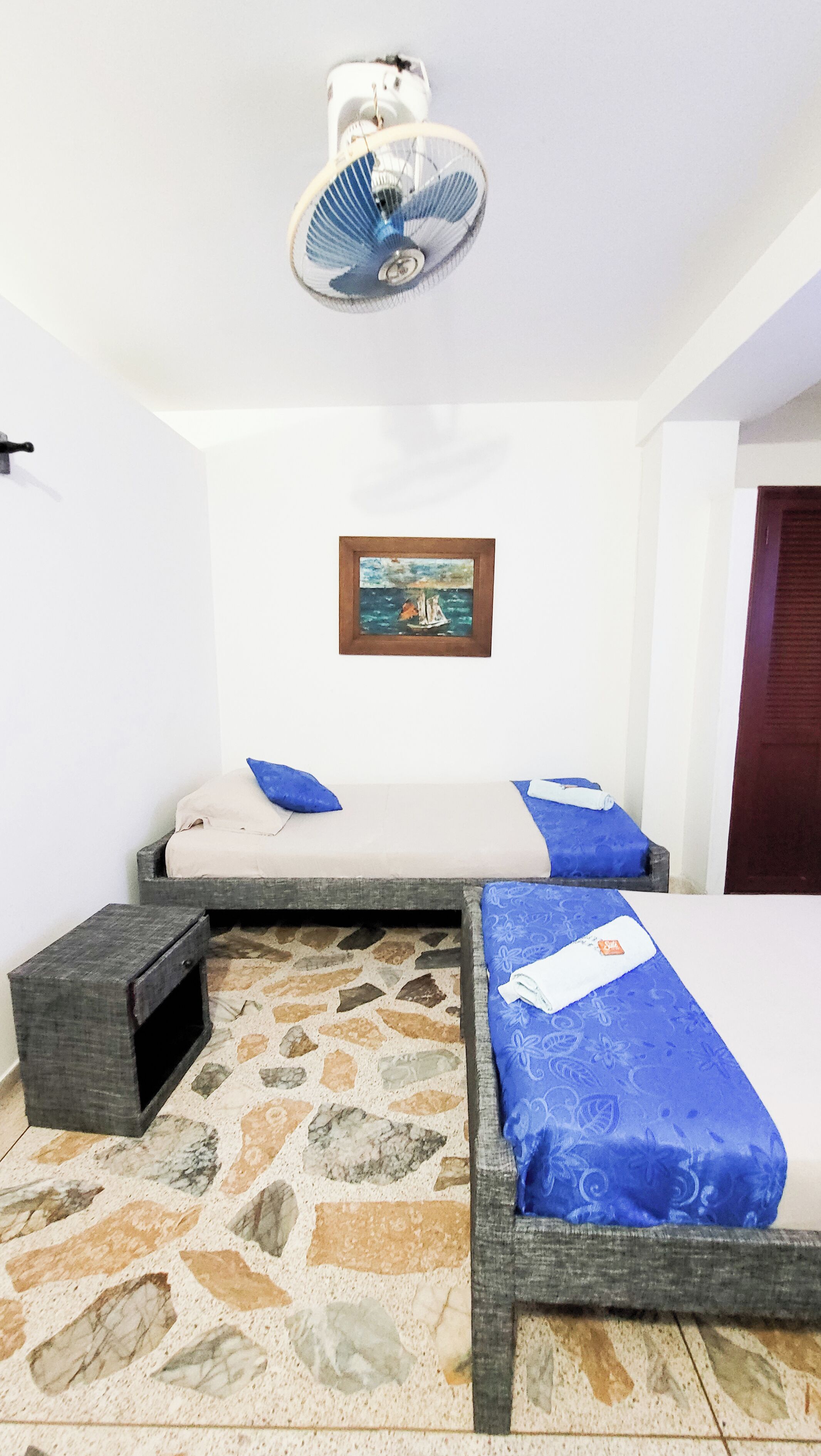 A room with two beds, a small table, a fan, and a painting on the wall.