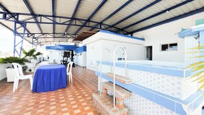 An outdoor patio with a blue table and chairs, a pool area with steps, and a covered roof structure.