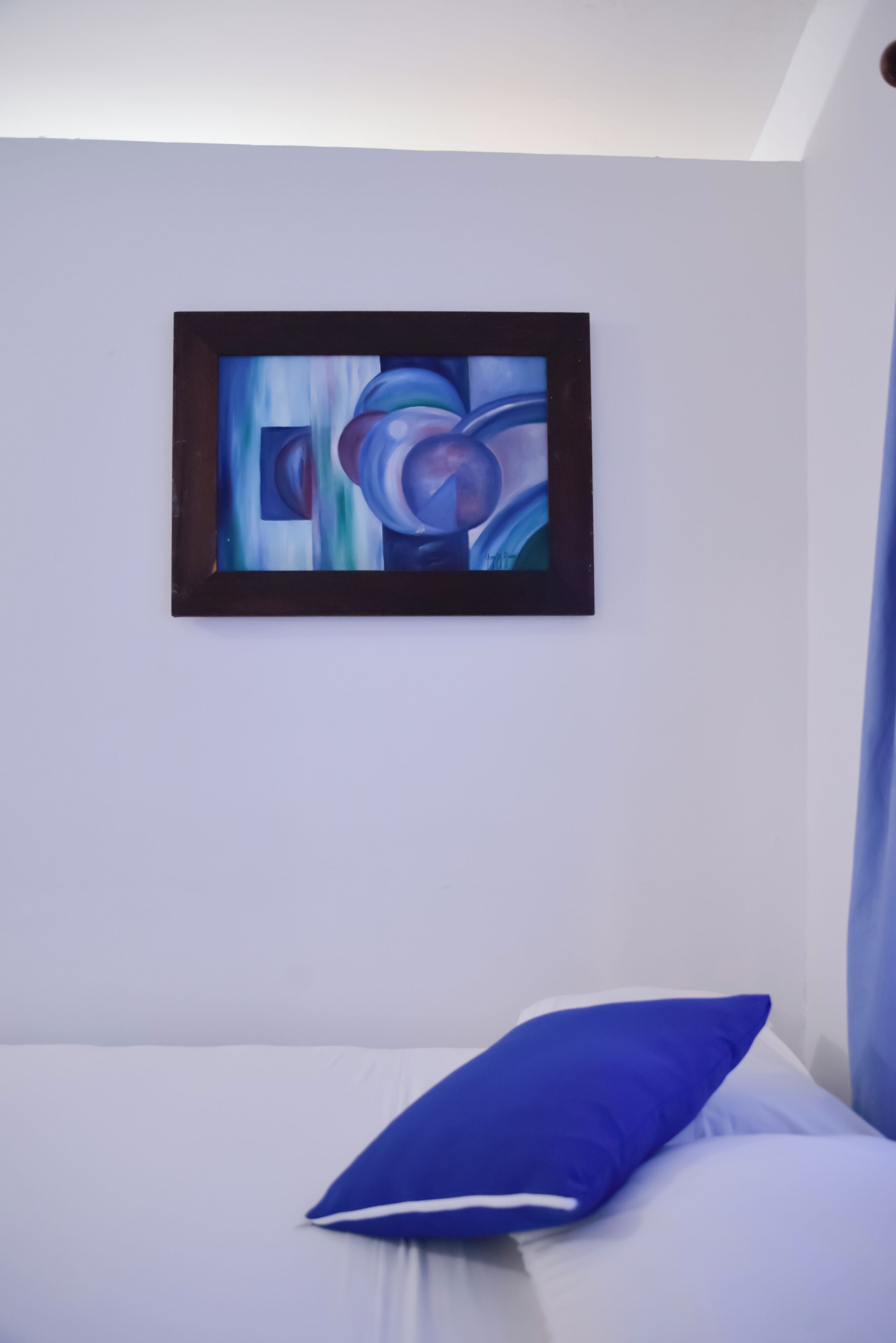 A bedroom with a bed, a blue pillow, and a framed abstract painting on the wall.