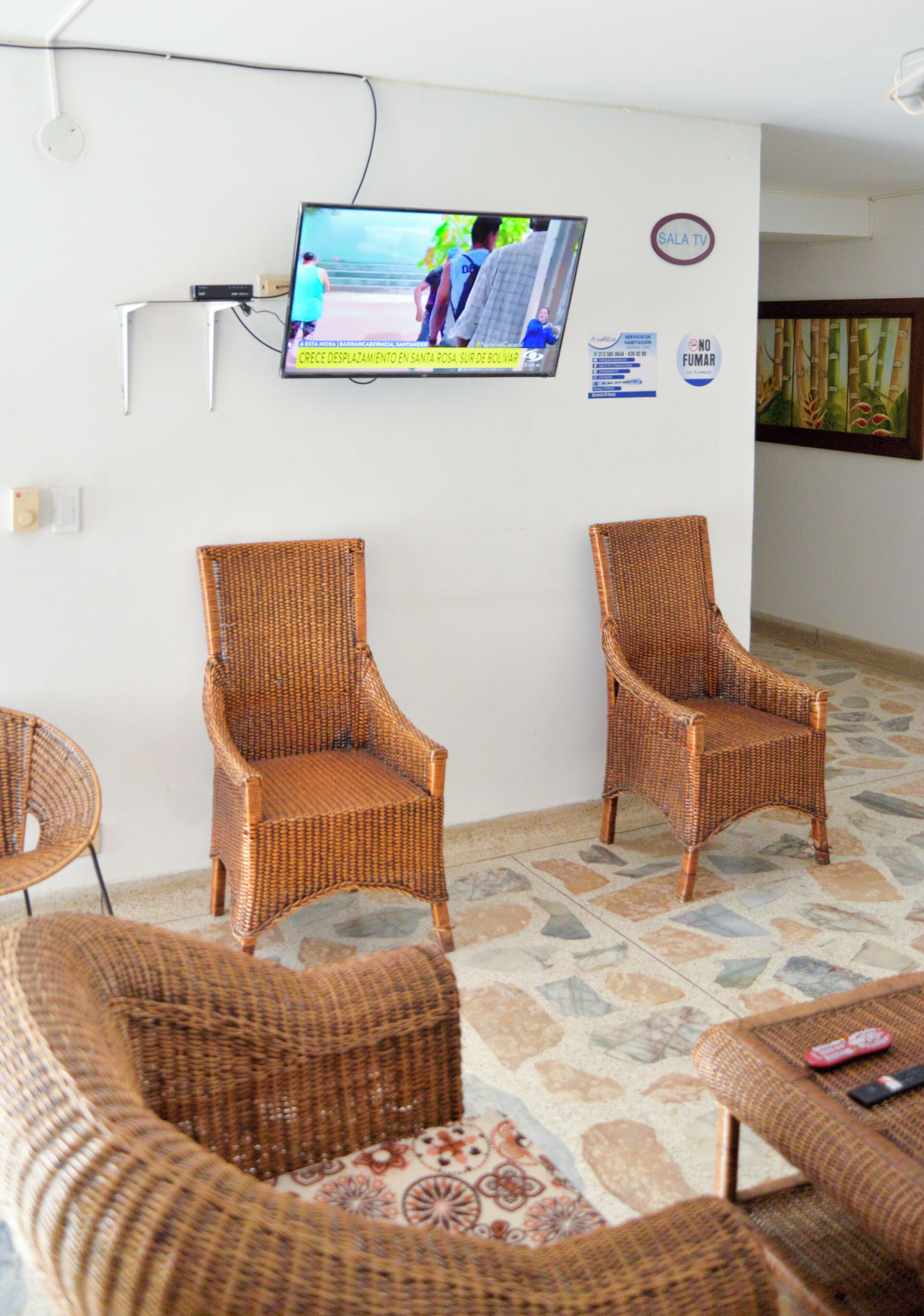 Una sala de estar con muebles de mimbre, un televisor de pantalla plana montado en la pared y suelo de baldosas.