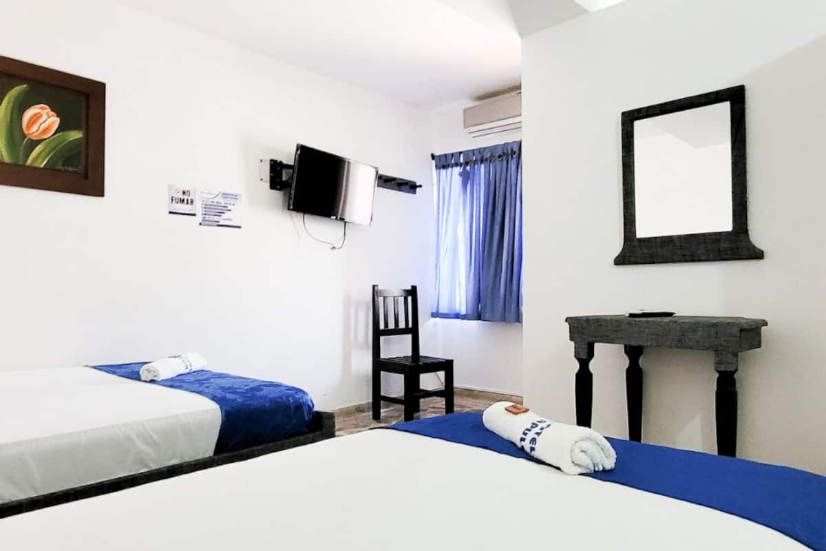Habitación de hotel con dos camas, una silla, una mesita, televisor y ventana con cortinas.