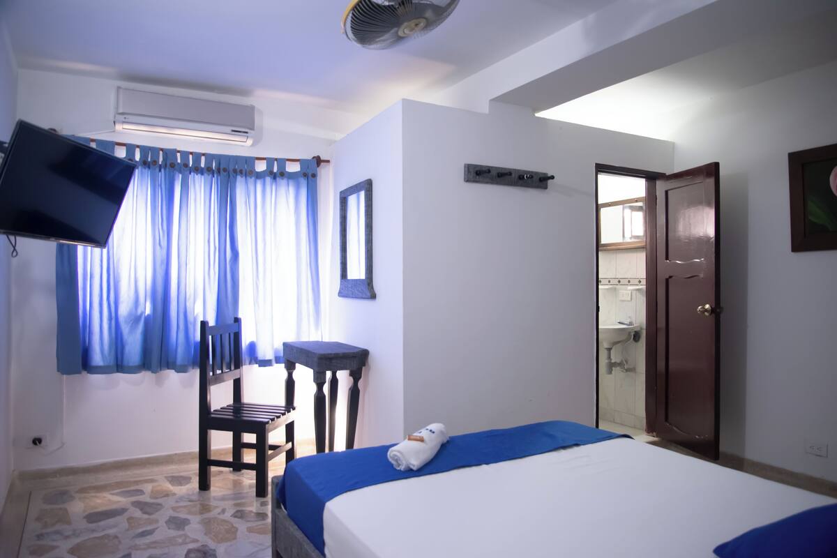 Habitación de hotel con cama, escritorio, silla, televisor y baño visible a través de una puerta abierta, y piso con diseño de piedra.