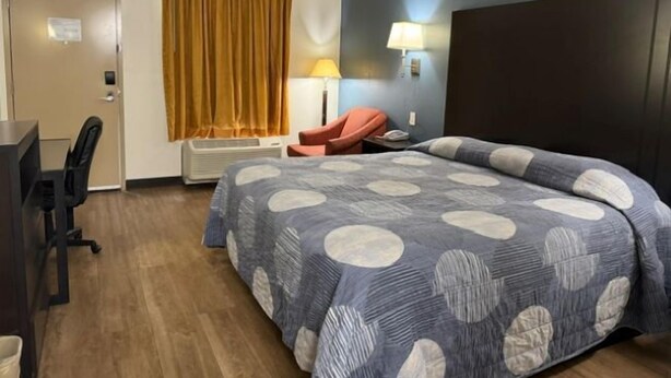 A hotel room with a bed, a desk, a chair, a lamp, and a window with curtains. - Hilltop Inn (Conley)
