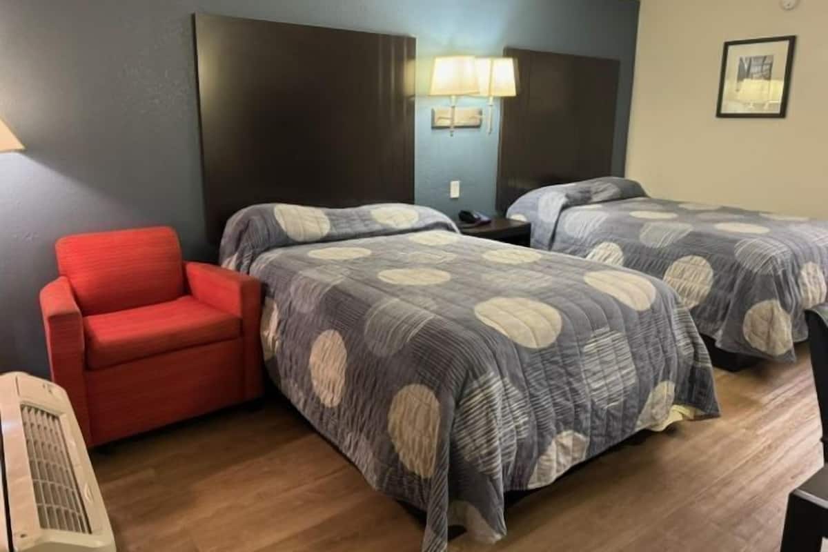 A hotel room with two beds, a red armchair, a wall-mounted lamp, and a framed picture on the wall.