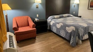 A hotel room with a bed, a chair, a nightstand, a lamp, and a heater. - Hilltop Inn (Conley)