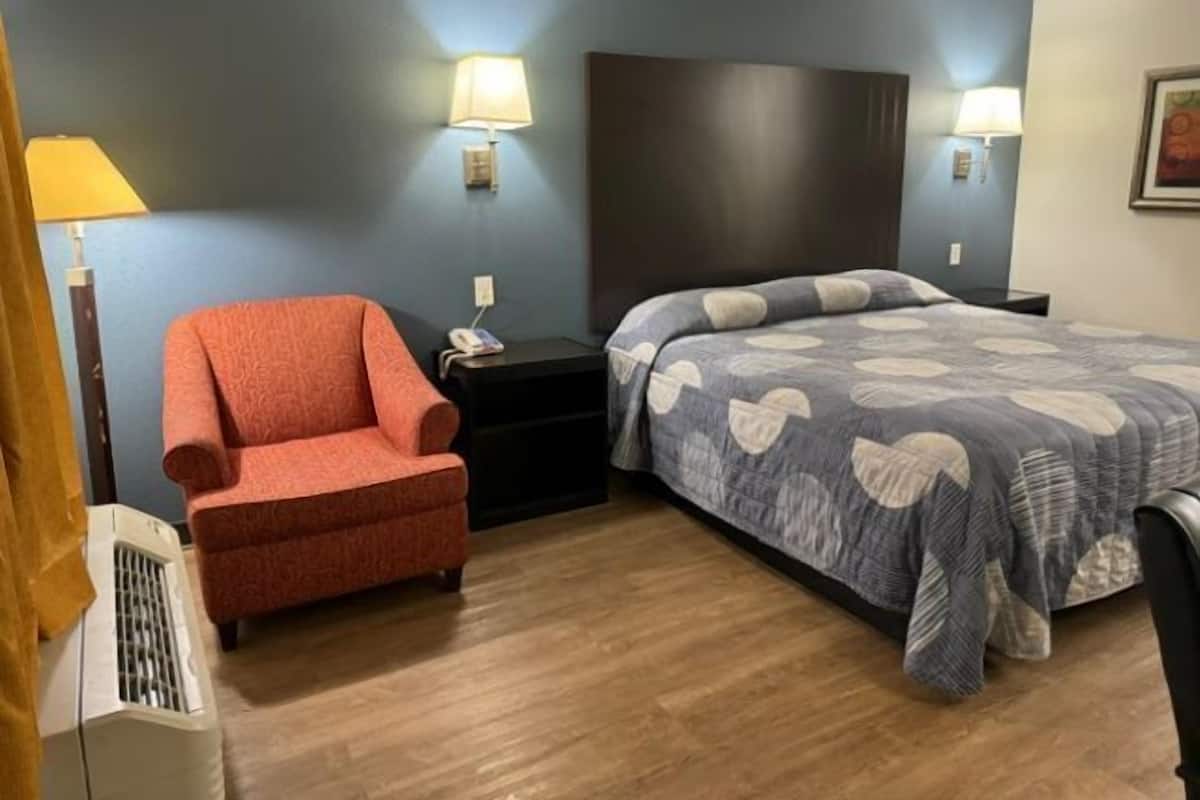 A hotel room with a bed, a chair, a nightstand, a lamp, and a heater.