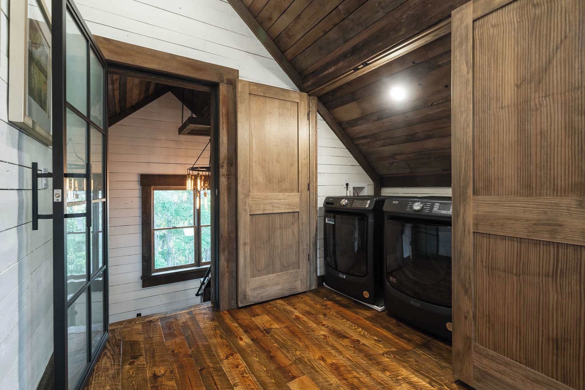 A laundry room with wooden walls and floors, a washing machine, and a window with a view of greenery.