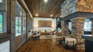 A spacious living area with a stone fireplace, wooden flooring, and large windows. - Stag's Manor|Stunning Mtn Views|Game Room|Hot Tub (Morganton)