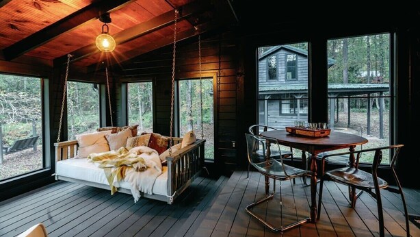L’intérieur chaleureux d’un chalet avec un lit-hamac en bois, une table à manger avec des chaises et de grandes fenêtres offrant une vue sur la forêt environnante.