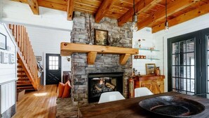 Un intérieur rustique avec une cheminée en pierre, des poutres en bois, un coin repas agrémenté d’un grand saladier et un escalier.