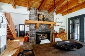 A rustic interior with a stone fireplace, wooden beams, a dining table with a large bowl, and a staircase. - Holden Haus | Creekside Cabin + Hot Tub + Dogs (Ellijay)