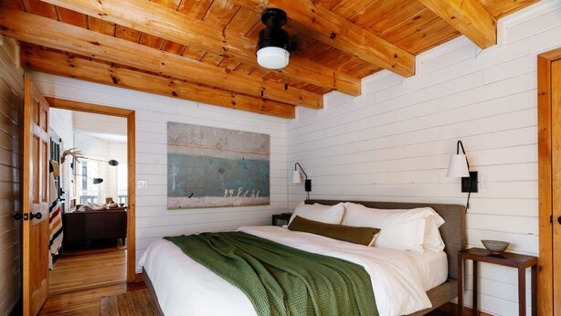 Een slaapkamer met een houten plafond, een bed met wit beddengoed, een groene plaid en een schilderij aan de muur.