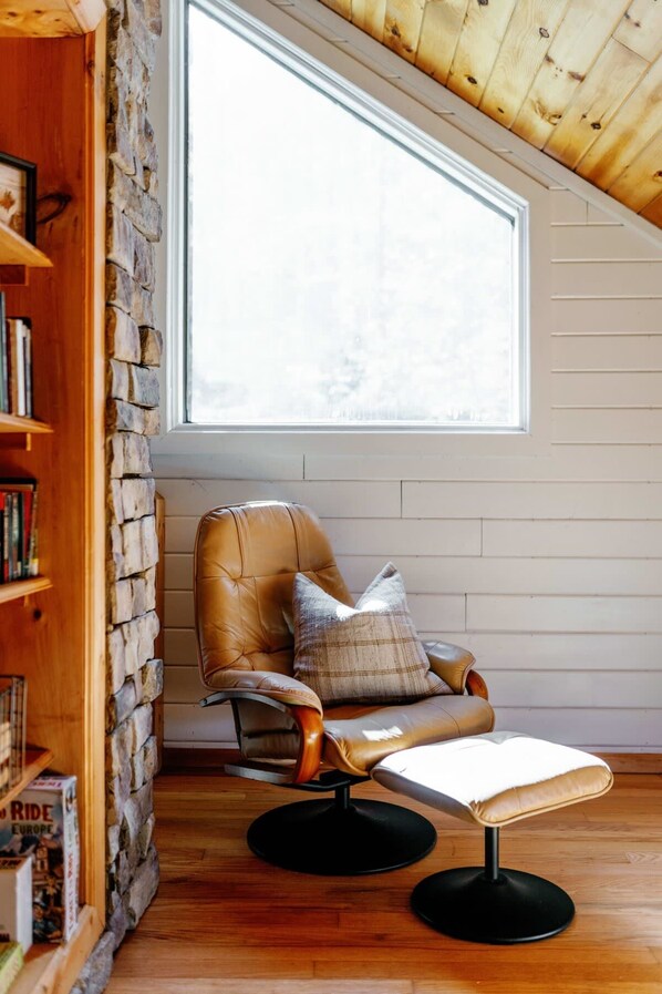 A cozy reading nook with a leather armchair, a small table, and a bookshelf against a stone wall, under a sloped ceiling with a large window.