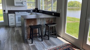 A modern kitchen with a large island, bar stools, and a view of a lake through the windows. - Rock River Retreat Boathouse Bungalow...Relax, Refresh, and Enjoy this Zen Den (Horicon)