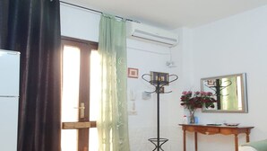 A room with a door, a window with curtains, a radiator, a mirror, a small table with a vase of flowers, and a patterned floor. - Apartment in Matera near Historic Center (Matera)