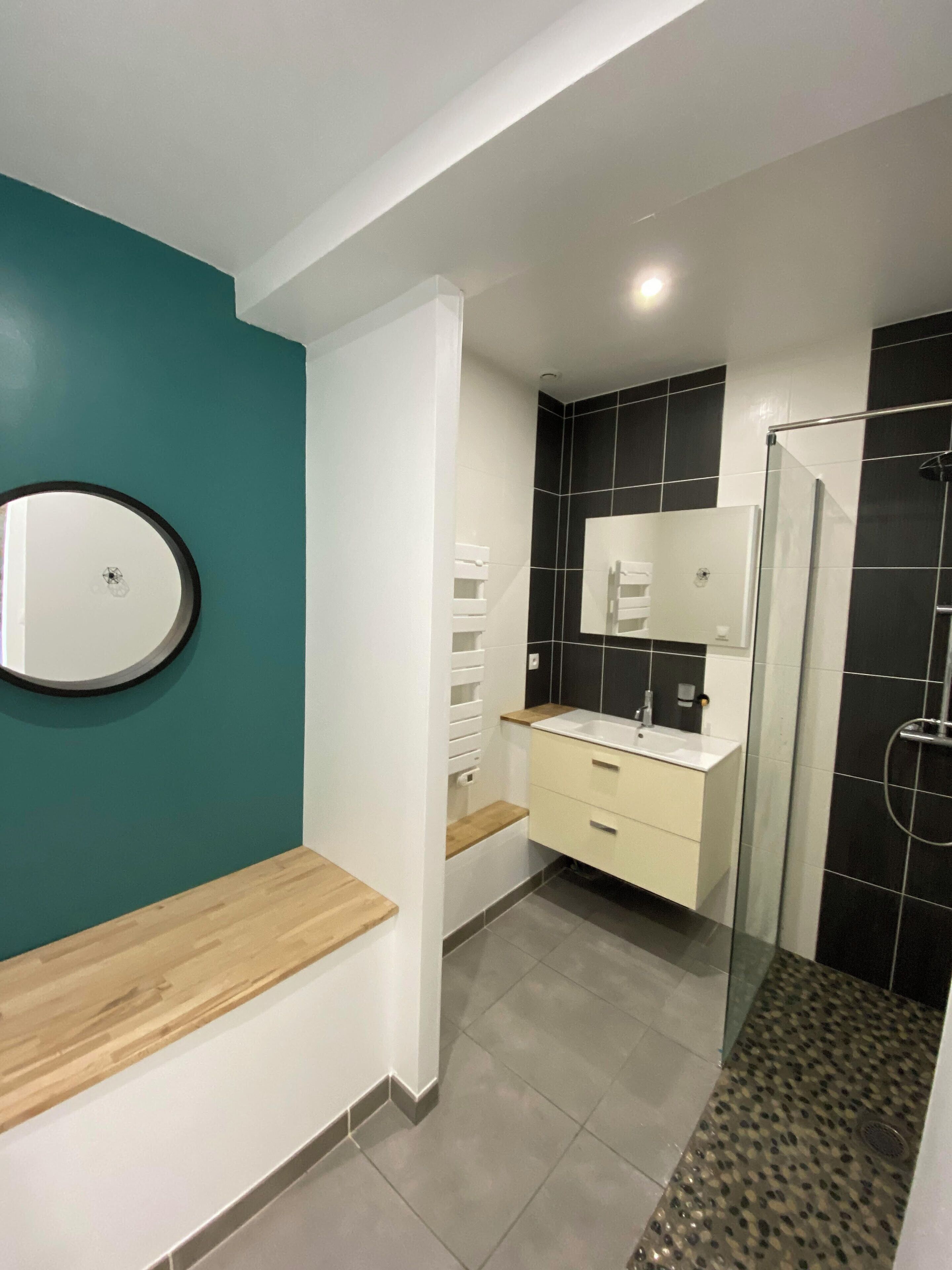 Une salle de bain moderne avec un mur d’accent de couleur turquoise, un miroir rond, un meuble-lavabo blanc et une cabine de douche en verre.