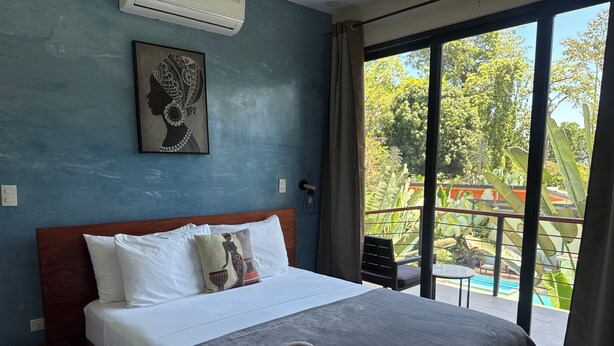 Una habitación de hotel con cama, ventana con vista a la vegetación y ventilador de techo.