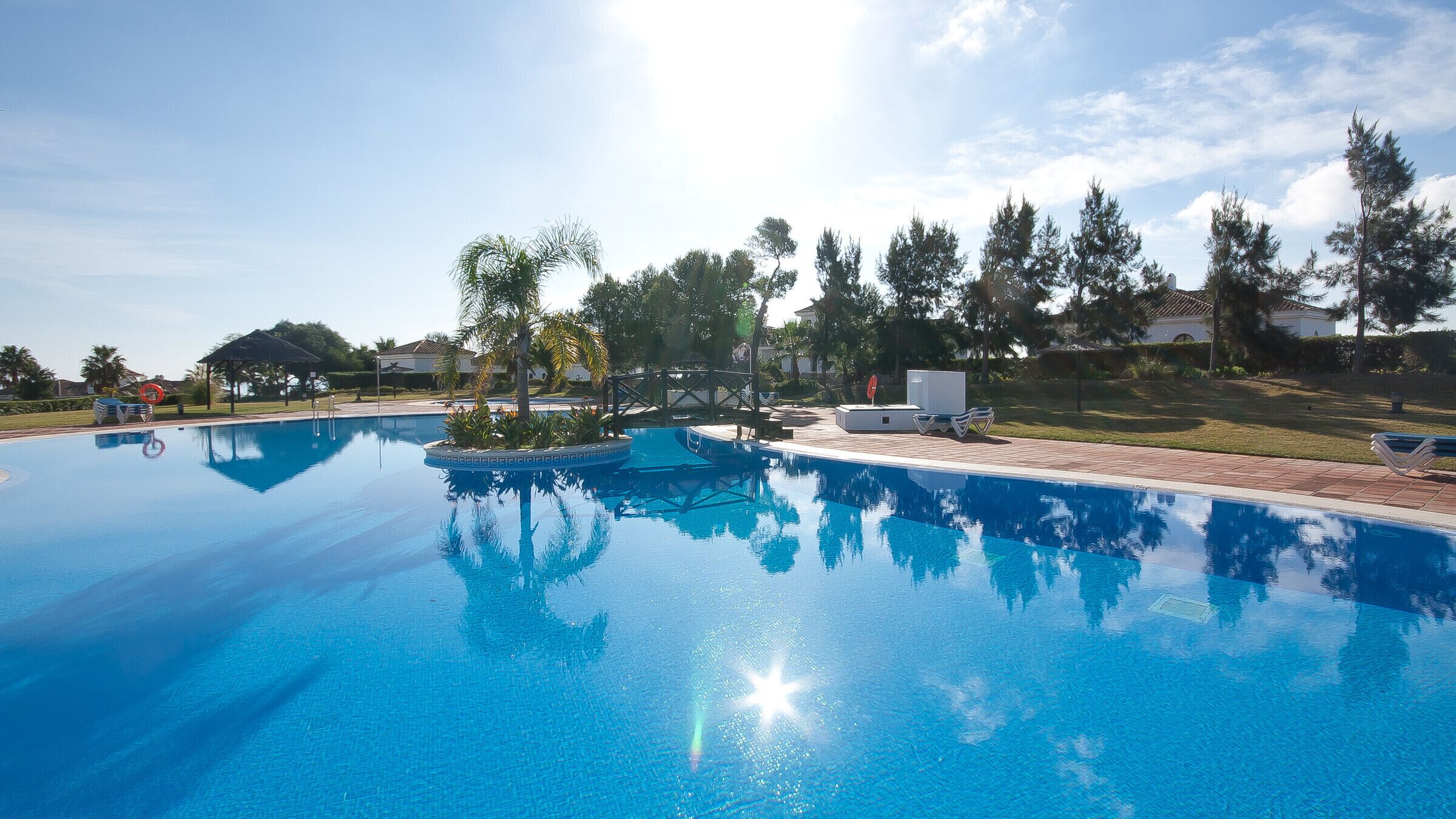 A clear blue swimming pool with a sun reflecting on the water, surrounded by palm trees and lounge chairs.