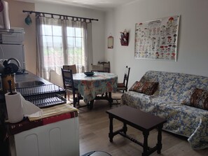 Uma sala de estar com sofá, mesa de jantar com cadeiras, área de cozinha com pia e eletrodomésticos, e um cartaz na parede.