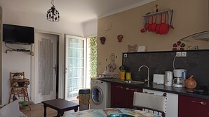 Een kleine keuken met een wasmachine, een spoelbak en een koffiezetapparaat. Er staat een eettafel met een tafelkleed met een wereldkaart erop en een kom.