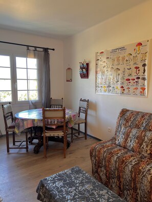 A dining area with a round table and chairs, a sofa, a poster of mushrooms, and a window with curtains.