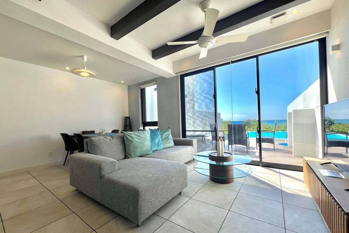 Una sala de estar moderna con un sofá seccional gris, una mesa de centro de vidrio y un amplio ventanal corredizo que da a una zona de piscina.