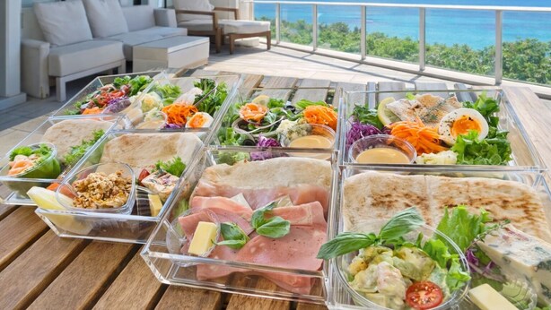 Un bufé con diversas opciones de comida, como ensaladas, sándwiches y carnes, sobre una terraza de madera con vista al océano.