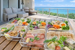 Un buffet proposant une variété de plats, notamment des salades, des sandwichs et des viandes, installé sur une terrasse en bois avec vue sur l’océan.