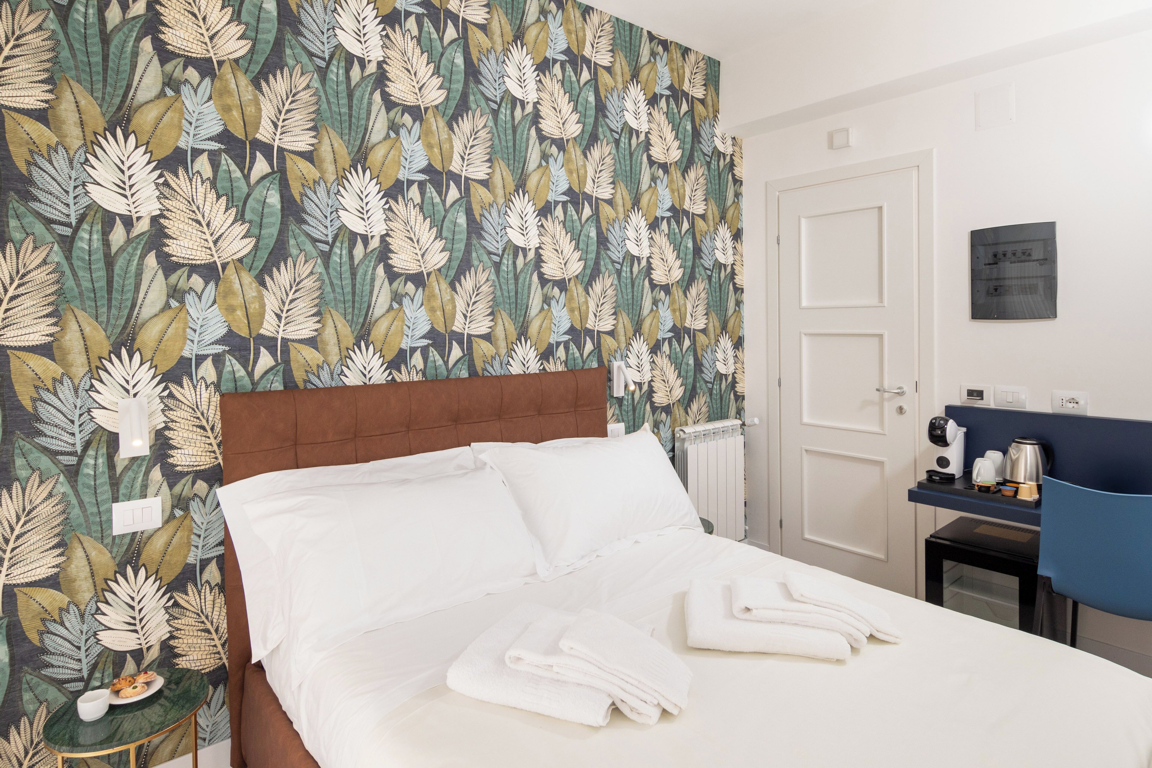 Een hotelkamer met een bed, witte lakens, een bruin hoofdeinde, een muur met een bladerpatroon, een klein tafeltje met een kopje en bord, en een blauwe stoel.