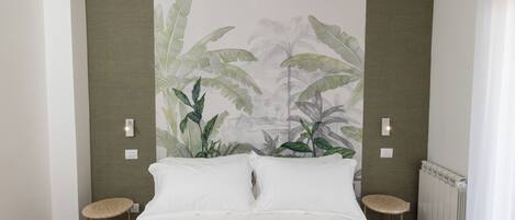 A bedroom with a bed, two bedside tables, a radiator, and a wall with a tropical plant mural.