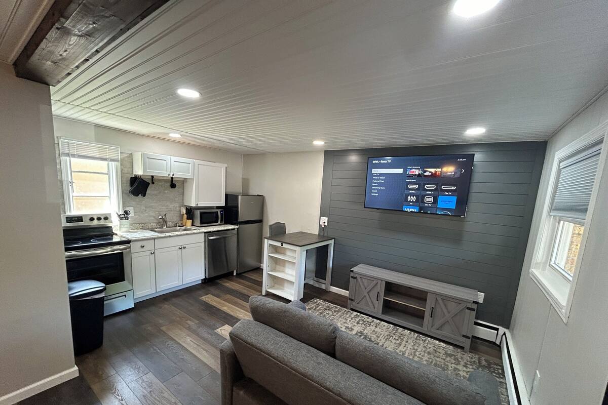 A compact living space with a kitchenette, a flat-screen TV mounted on the wall, and a grey sofa.