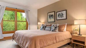 A bedroom with a large bed, two bedside tables, a window with curtains, and two framed artworks on the wall.