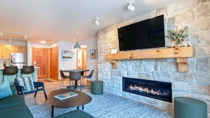A modern living room with a stone fireplace, a flat-screen TV mounted on a wooden shelf, and a dining area in the background.
